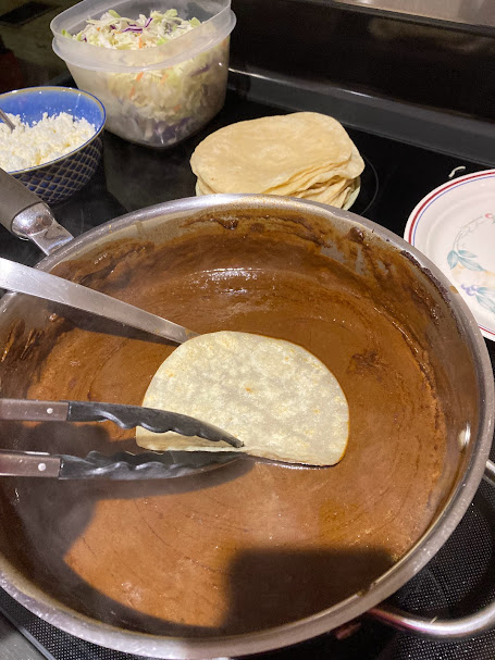 tortilla being dipped into the hot mole, with other ingredients on stove in background