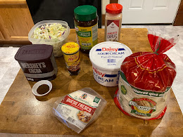 Container of cabbage, cocoa powder, jarred mole, arbol powder, chicken bouillon, garlic powder, tortillas, and sour cream on a chopping block in the kitchen