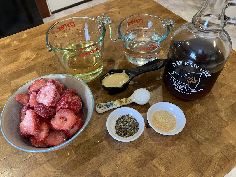 Ingredients for the Strawberry Maple Vinaigrette Salad Dressing.  Frozen strawberries in a bowl, oil and vinegar in measuring cups, dijon mustard in a cup, salt in a spoon, as well as a bottle of Pure New York Maple Syrup and two small dishes with pepper and garlic